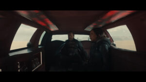 The interior of a dark red 1990s Lincoln Town Car limo as seen in the Marvel Studios Film Thunderbolts*. The character John Walker can be seen sitting in the back seat with the character "Ghost" sitting on the right side of the frame. Above them both is red mood lighting panels mounted in the ceiling of the limo.