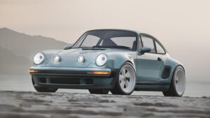 Front ¾ showing carbon fibre bodywork in Celeste Passalacqua, touring-focused front fascia with splitter, 18” centre lock wheels and rear fender cooling intake. High-performance auxiliary driving lights are specified and shown in the raised position.